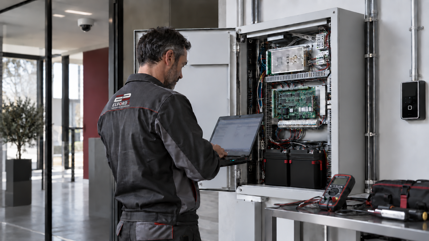 Immagine illustrativa di un tecnico con divisa Elford2 durante la diagnosi di un sistema di sicurezza, con computer portatile accanto alla centrale aperta.